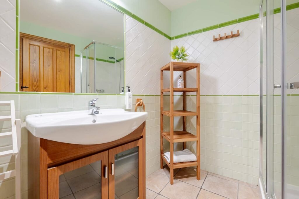 Aseo en tonos verdes de Casa Rural Castalla con ducha acristalada, lavabo y estantería de madera